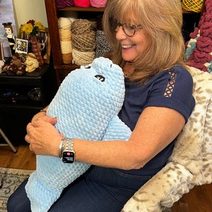 May include: A woman is holding a blue crocheted stuffed animal that resembles a seal. The stuffed animal has black eyes and a white mouth. The woman is wearing a blue shirt, blue jeans, and a silver watch.