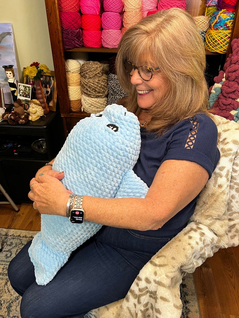 May include: A woman is holding a blue crocheted stuffed animal that resembles a seal. The stuffed animal has black eyes and a white mouth. The woman is wearing a blue shirt, blue jeans, and a silver watch.