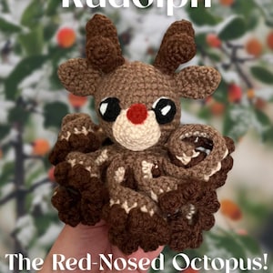 Op de afbeelding: Een gehaakte Rudolph, het rendier met de rode neus, octopus met een rode neus en bruine en witte tentakels. De octopus wordt in een hand gehouden tegen een wazige achtergrond van groene bladeren en rode bessen.