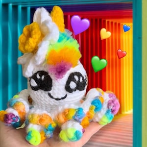 以下が含まれることがあります： 手作りの白いかぎ針編みのタコ、虹色のアクセント付き。タコは大きな黒い目、笑顔の口、カラフルな触手を持っています。タコは虹色のたてがみがあり、黄色と虹色のアクセントで飾られています。カラフルなハートの絵文字が背景にあります。