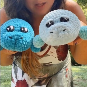 May include: Two crocheted plush toys in the shape of a manatee. One is blue and the other is light blue. Both have black eyes and a white nose.