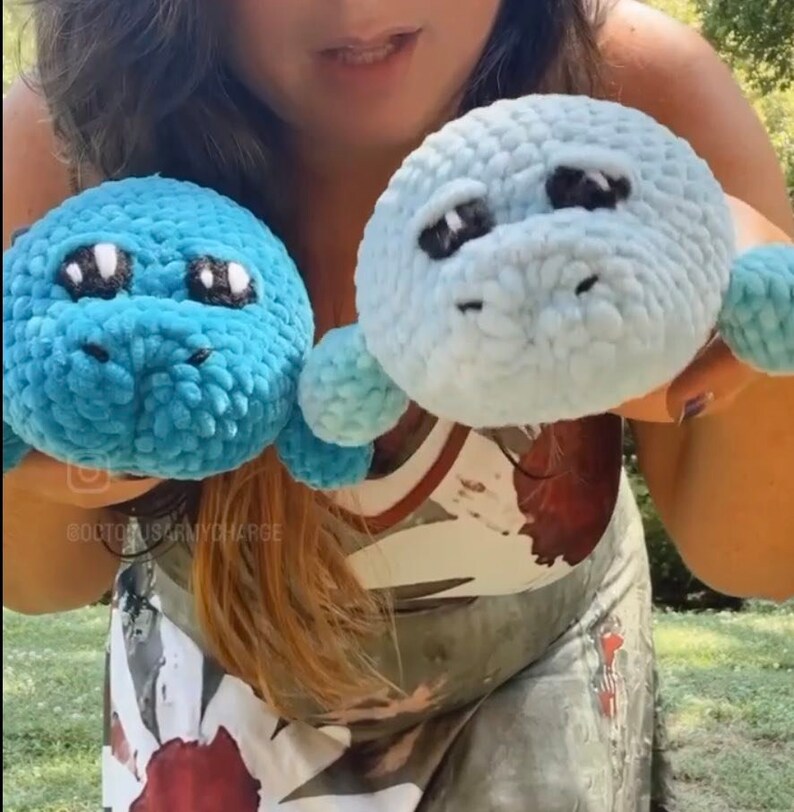 May include: Two crocheted plush toys in the shape of a manatee. One is blue and the other is light blue. Both have black eyes and a white nose.