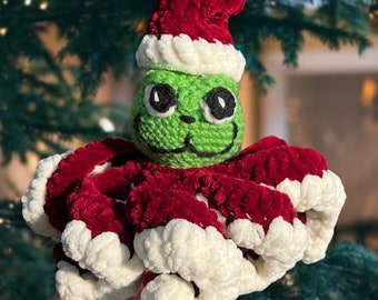 Könnte beinhalten: Ein grüner gehäkelter Oktopus, der einen roten Samt-Nikolaus-Hut mit weißem Besatz trägt. Der Oktopus hat schwarze Augen und ein weißes Lächeln.
