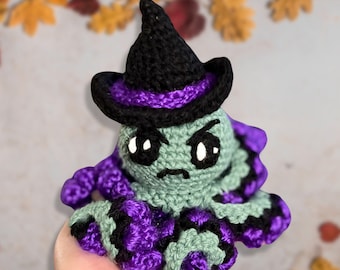 May include: A crocheted octopus wearing a black witch hat with a purple band. The octopus has a green body with black and purple tentacles and has a grumpy expression on its face.
