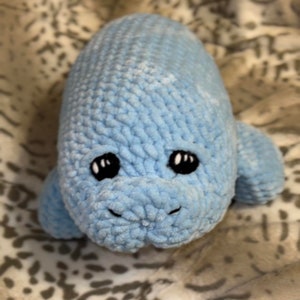 May include: A blue crocheted stuffed manatee toy with black eyes. The toy is lying on a white and grey patterned blanket.