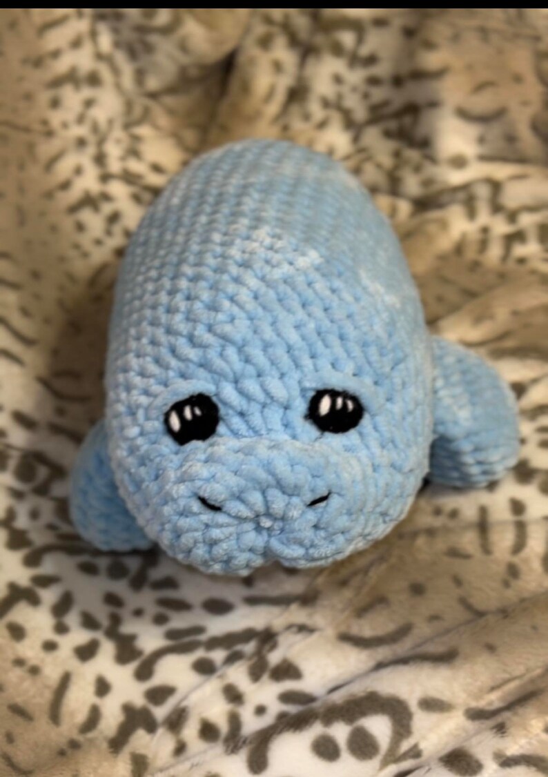 May include: A blue crocheted stuffed manatee toy with black eyes. The toy is lying on a white and grey patterned blanket.