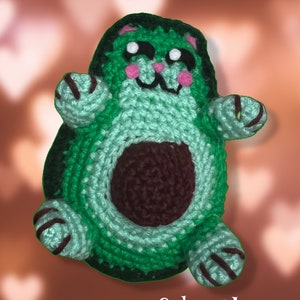 May include: Green crocheted avocado cat toy with brown details. The cat has a cute face with pink cheeks and a brown nose. The toy is made with a soft yarn and is perfect for cuddling.
