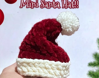 May include: A red and white crocheted Santa hat with a white pom-pom on top. The hat is small and is being held in a hand. The text "No-Sew Mini Santa Hat!!" is written above the hat. The text "OctopusArmy Crochet" is written below the hat.