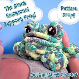 Könnte beinhalten: Ein grüner und blauer gehäkelter Frosch mit schwarzen Augen. Der Frosch sitzt auf dem Schoß einer Person. Der Text "The Giant Emotional Support Frog!" befindet sich in einer Sprechblase über dem Frosch. Der Text "Pattern Drop!" befindet sich in einer Sprechblase rechts neben dem Frosch. Der Text "OctopusArmy Crochet" befindet sich am unteren Rand des Bildes.