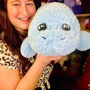 May include: A woman is holding a light blue crocheted stuffed animal that resembles a manatee. The stuffed animal has black eyes and a small mouth.