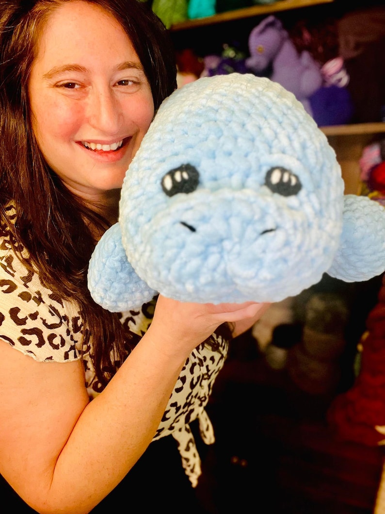 May include: A woman is holding a light blue crocheted stuffed animal that resembles a manatee. The stuffed animal has black eyes and a small mouth.