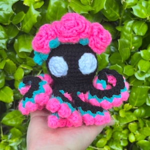 May include: A crocheted octopus with pink and teal tentacles and a pink flower crown. The octopus has black eyes and a black body.
