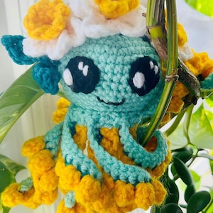 May include: A crocheted octopus with a teal body and yellow tentacles. The octopus is wearing a white flower crown with yellow centers.