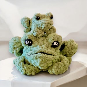 May include: Two crocheted green frog plushies. The smaller frog is sitting on top of the larger frog. Both frogs have black eyes.