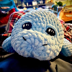 May include: A light blue crocheted stuffed animal shaped like a manatee with black eyes. The manatee is lying on a blue surface.