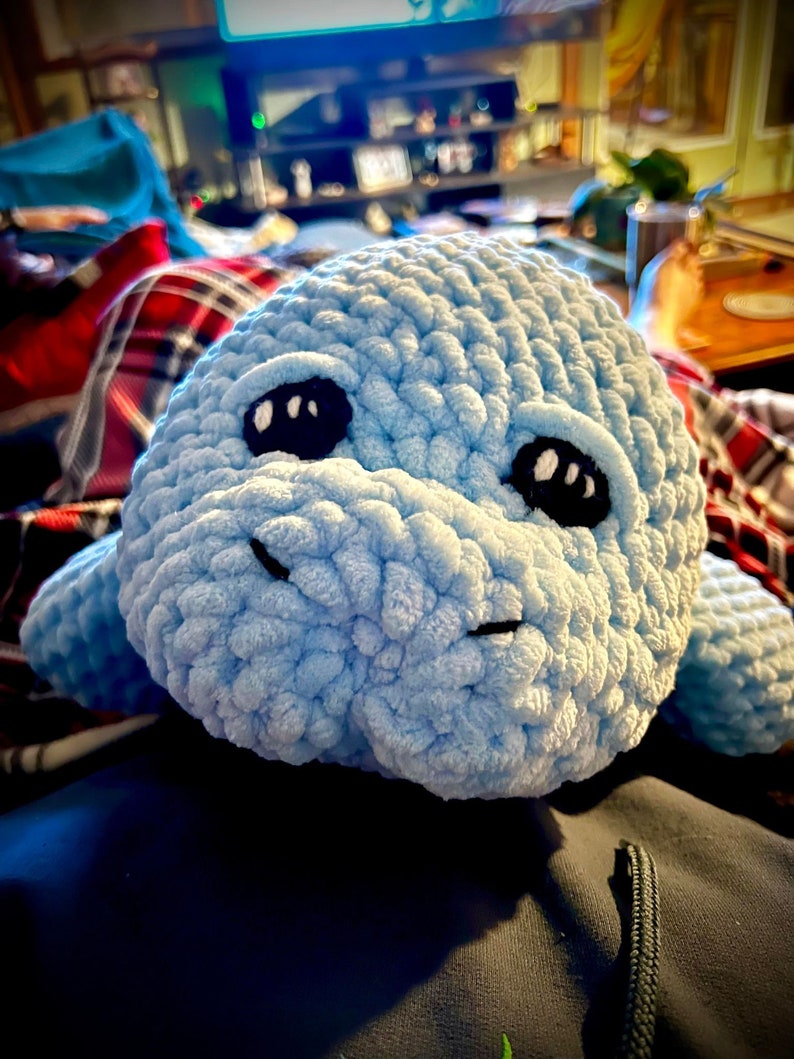 May include: A light blue crocheted stuffed animal shaped like a manatee with black eyes. The manatee is lying on a blue surface.