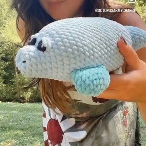 May include: A person is holding a light blue crocheted sea lion plush toy. The toy has a light blue fin and black eyes. The person is wearing a white and red patterned shirt.