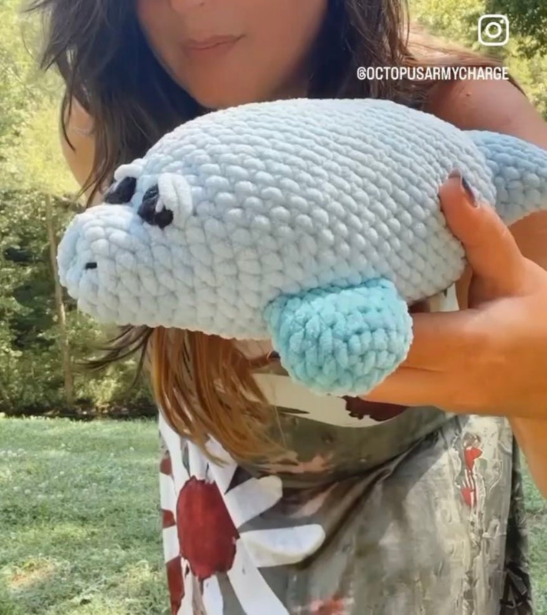 May include: A person is holding a light blue crocheted sea lion plush toy. The toy has a light blue fin and black eyes. The person is wearing a white and red patterned shirt.