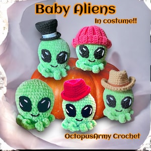 May include: Five crocheted baby alien toys wearing different costumes. The aliens are green with large black eyes and a smiling mouth. They are sitting on a pumpkin. The text "Baby Aliens In costume!!" is at the top of the image. The text "OctopusArmy Crochet" is at the bottom of the image.