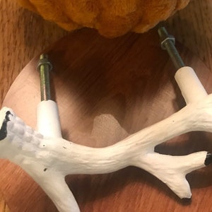 May include: A white ceramic antler-shaped cabinet knob with a brown wooden base. The knob is attached to a screw. The knob is sitting on a wooden surface.
