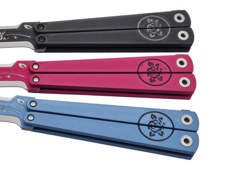 Op de afbeelding: Drie vlindermessen in zwart, roze en blauw. Elk mes heeft een rozenembleem op het handvat en een bloemmotief op het lemmet. De handvatten zijn rechthoekig met afgeronde randen en hebben twee gaten voor de schroeven.
