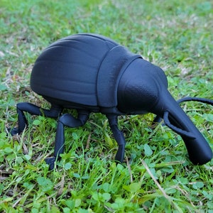 Könnte beinhalten: Eine schwarze, 3D-gedruckte Käferfigur mit langem Rüssel und segmentiertem Körper. Das Insekt ist auf grünem Gras platziert und zeigt seine detaillierten Beine und Fühler. Der Körper des Käfers hat eine strukturierte Oberfläche.