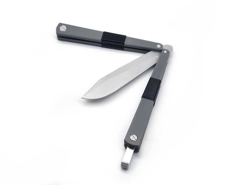 May include: A gray and black butterfly knife with a silver blade, partially open. The handle is gray with black rubber grips. The blade is angled, showing the sharp edge. The knife is on a white background.