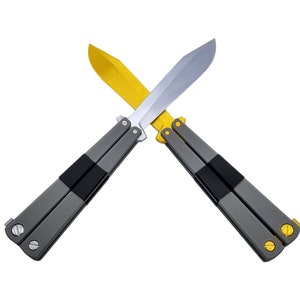 May include: Two butterfly knives crossed against a white background. One knife has a gold blade, the other a silver blade. Both knives have gray handles with black rubber grips and gold accents.