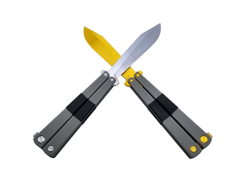 May include: Two butterfly knives crossed against a white background. One knife has a gold blade, the other a silver blade. Both knives have gray handles with black rubber grips and gold accents.