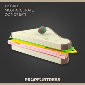 May include: A 1:1 scale model of a sandwich with layers of pink, yellow, and green fillings between white bread slices. A green olive with a red dot sits atop the sandwich. The text "1:1 SCALE MOST ACCURATE DO NOT EAT!" is visible.