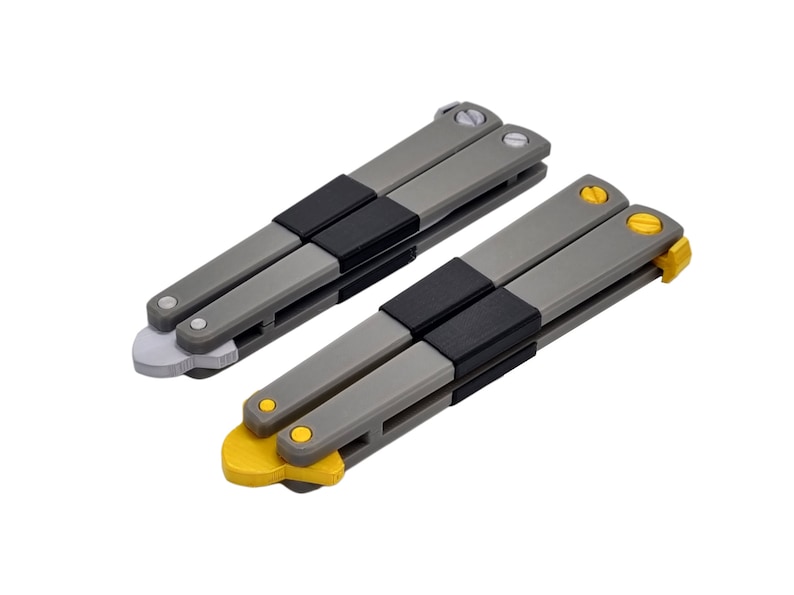 May include: Two butterfly knives with gray handles, black center grips, and silver or yellow accents. The blades are folded, showcasing the design and color variations. The knives are set against a white background.