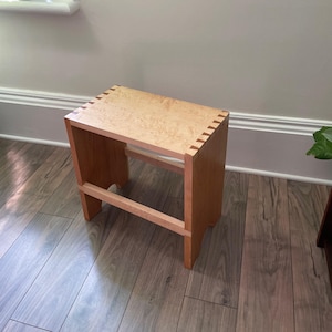 Peut inclure: Un petit tabouret en bois avec une finition en bois naturel. Le tabouret a un design simple avec un dessus rectangulaire et quatre pieds. Le dessus du tabouret est en bois clair avec un motif de grain naturel. Les pieds sont en bois plus foncé avec une finition lisse.