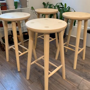 May include: Four wooden bar stools with round seats and four legs each. The stools are made of light-colored wood and have a simple, minimalist design. The seats have small, darker wood accents.