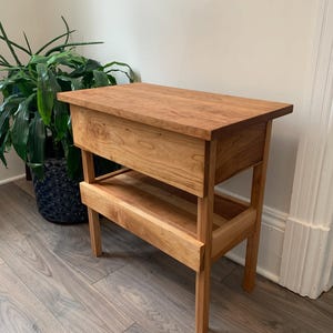 Peut inclure: Une table d'appoint en bois avec une étagère et un tiroir. La table est en bois brun clair avec une finition naturelle. La table est posée sur un sol en bois brun clair.
