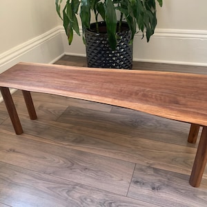 May include: A natural wood bench with a live edge top and two legs. The bench is made of walnut wood and has a dark brown colour.