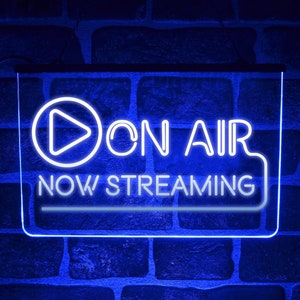 Peut inclure: Un panneau lumineux bleu néon avec un bouton de lecture blanc et le texte "ON AIR NOW STREAMING".