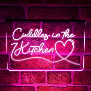 May include: Pink neon sign with white lettering that reads "Cuddles in the Kitchen" with a pink heart.