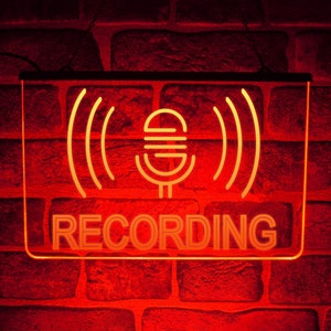 May include: A neon sign with a microphone and sound waves, and the word "RECORDING" in yellow neon letters against a red brick wall background.