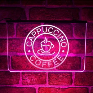 Op de afbeelding: Een roze neon-bord met een witte cirkel waarop "Cappuccino Coffee" staat, met een gestileerd beeld van een kopje koffie in de cirkel.