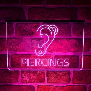 Może przedstawiać: Różowy neon z białym napisem "PIERCINGS" i zarysem ucha z zaznaczonymi miejscami przekłuć. Znak, wskazujący na usługi piercingu, jest umieszczony na tle czerwonej ceglanej ściany.