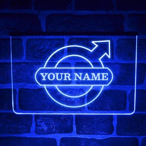 Peut inclure: Enseigne au néon bleue avec le texte "YOUR NAME" dans un rectangle blanc arrondi. Le signe présente un design circulaire avec une flèche pointant vers le haut. Monté sur un mur de briques bleues.