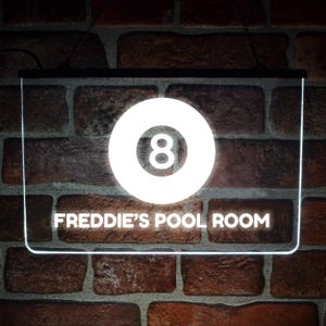 May include: A rectangular LED sign with a black frame featuring a glowing white graphic of an eight ball and the text FREDDIE'S POOL ROOM in white capital letters. The sign is illuminated against a dark red brick wall background.