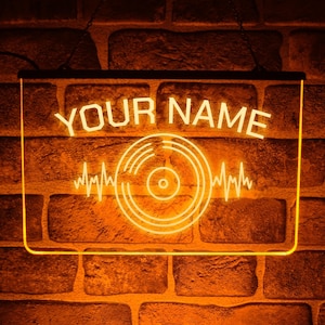 Könnte beinhalten: Ein rechteckiges LED-Schild mit leuchtend orangefarbenem Licht. Das Schild zeigt oben den Text YOUR NAME und darunter eine Grafik einer Schallplatte, die auf beiden Seiten von horizontalen Schallwellenlinien flankiert wird.