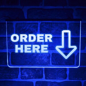 Op de afbeelding: Een blauw neonbord met de witte tekst "ORDER HERE" en een witte pijl naar beneden. Het bord is rechthoekig en bevestigd aan een donkere bakstenen muur.