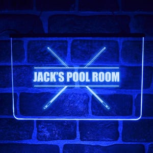 May include: A rectangular LED sign with a bright blue glow featuring the text JACK'S POOL ROOM in capital letters. The design includes two crossed pool cues positioned behind the text, all illuminated against a dark brick wall background.