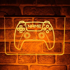 Può includere: Un'insegna da parete illuminata con un design a controller per videogiochi. Il contorno del controller si illumina con una calda luce arancione, con la parola "NAME" al centro. L'insegna è montata su un muro di mattoni.