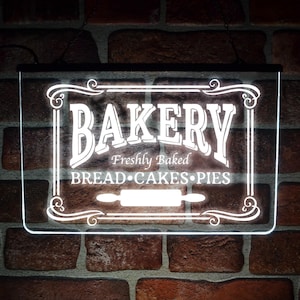 Op de afbeelding: Een rechthoekig, verlicht bord met het woord "BAKERY" in witte letters. Daaronder staat "Freshly Baked BREAD • CAKES • PIES". Een deegroller is in het midden geplaatst. Het bord is wit omlijst en hangt aan een bakstenen muur.