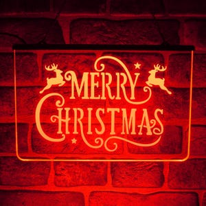 Może przedstawiać: Prostokątny podświetlany znak z napisem "MERRY CHRISTMAS" w ozdobnej czcionce, z sylwetkami reniferów. Znak emituje pomarańczową poświatę na tle czerwonej ceglanej ściany, tworząc świąteczną atmosferę.