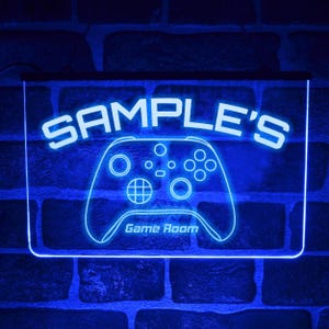 Könnte beinhalten: Blaues Neon-Schild mit dem Text "SAMPLE'S" und einer Controller-Umrisslinie. Die Worte "Game Room" stehen darunter. Das Schild ist an einer Backsteinmauer befestigt.