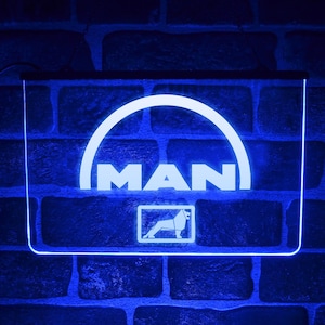 Può includere: Un'insegna a LED blu con la scritta "MAN" in bianco, posta su un muro di mattoni. L'insegna presenta un design ad arco bianco sopra il testo e un logo rettangolare sottostante. L'insegna è illuminata da una luce blu.